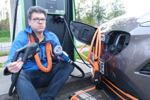 Bernd hat einen CCS und einen CHAdeMO Stecker in der Hand und ein AC Ladekabel um den Hals. An der Ladeluke des Autos hängt ein weiteres Ladekabel